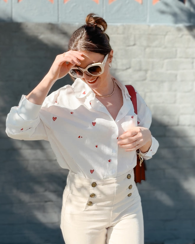 Woman wearing a white blouse with red heart patterns and white pants against a gray wall.