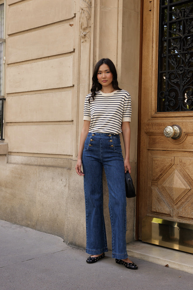 Woman in striped shirt and blue jeans standing against a building entrance. Jeans Victor Blue Maison Torrini Paris