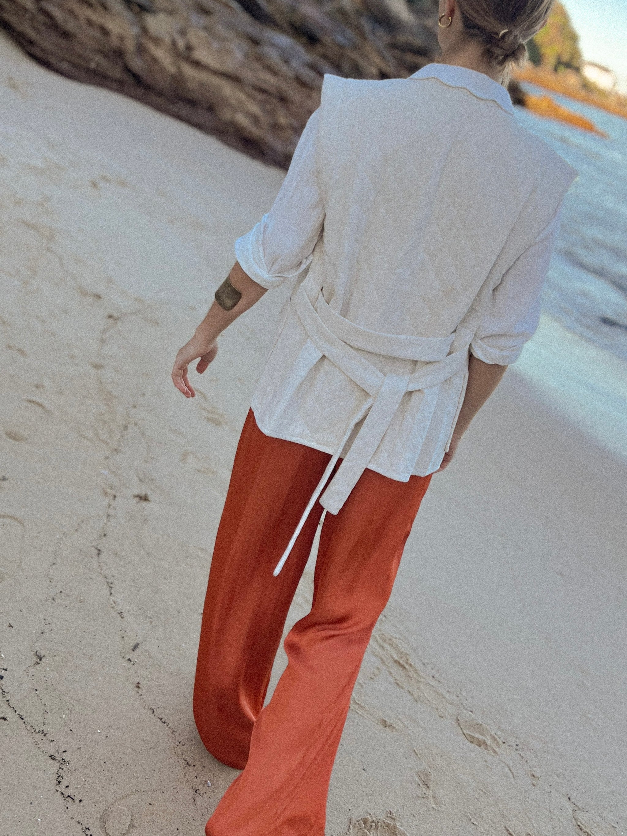 Maison Torrini Store, French labels, woman walking on the beach, wearing a terracotta satin pants, beige linen sleeveless jacket